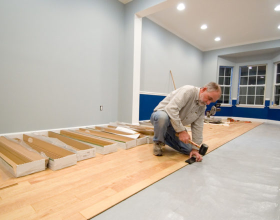 Man using a rubber mallet and tapping block to install the next row of laminate flooring. Adding to the do it yourself and renovation themes of the image are uncovered wall outlets and power tools in the background.