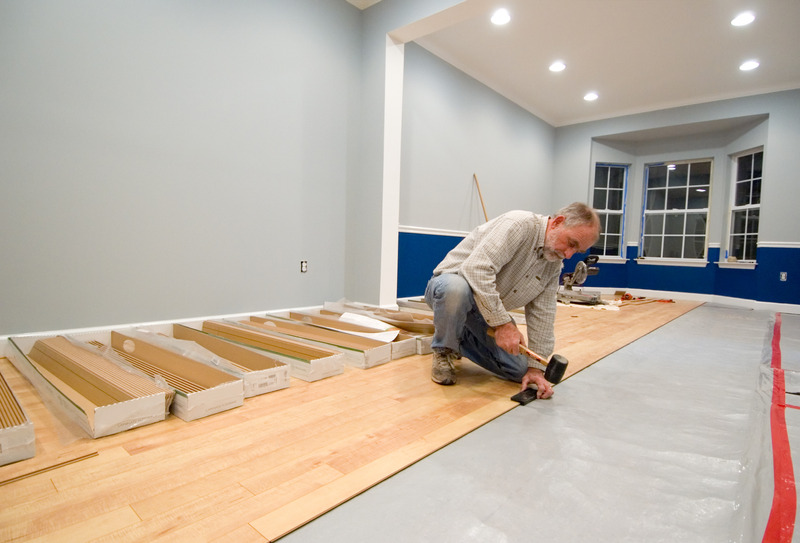 Man using a rubber mallet and tapping block to install the next row of laminate flooring. Adding to the do it yourself and renovation themes of the image are uncovered wall outlets and power tools in the background.